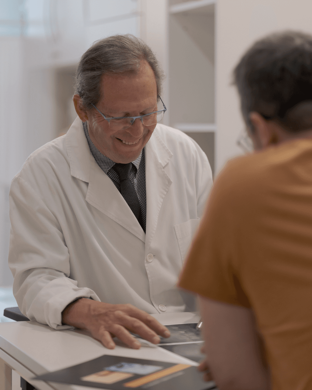 Médico sonriente con bata blanca y gafas revisando informes médicos con un paciente.