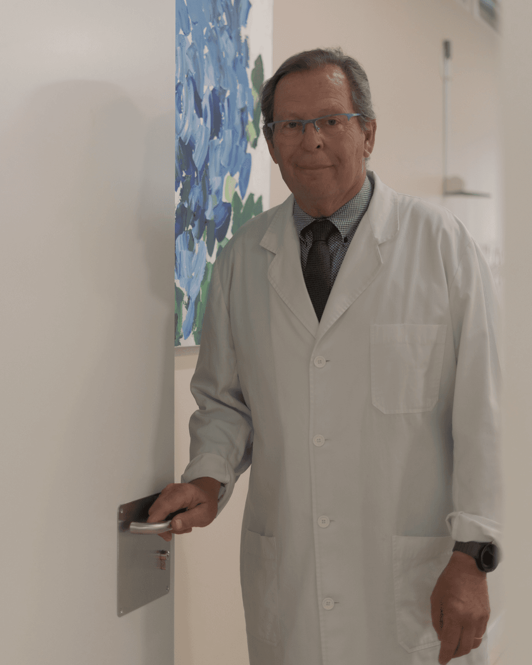 Médico con gafas y bata blanca abriendo la puerta de una consulta médica iluminada.