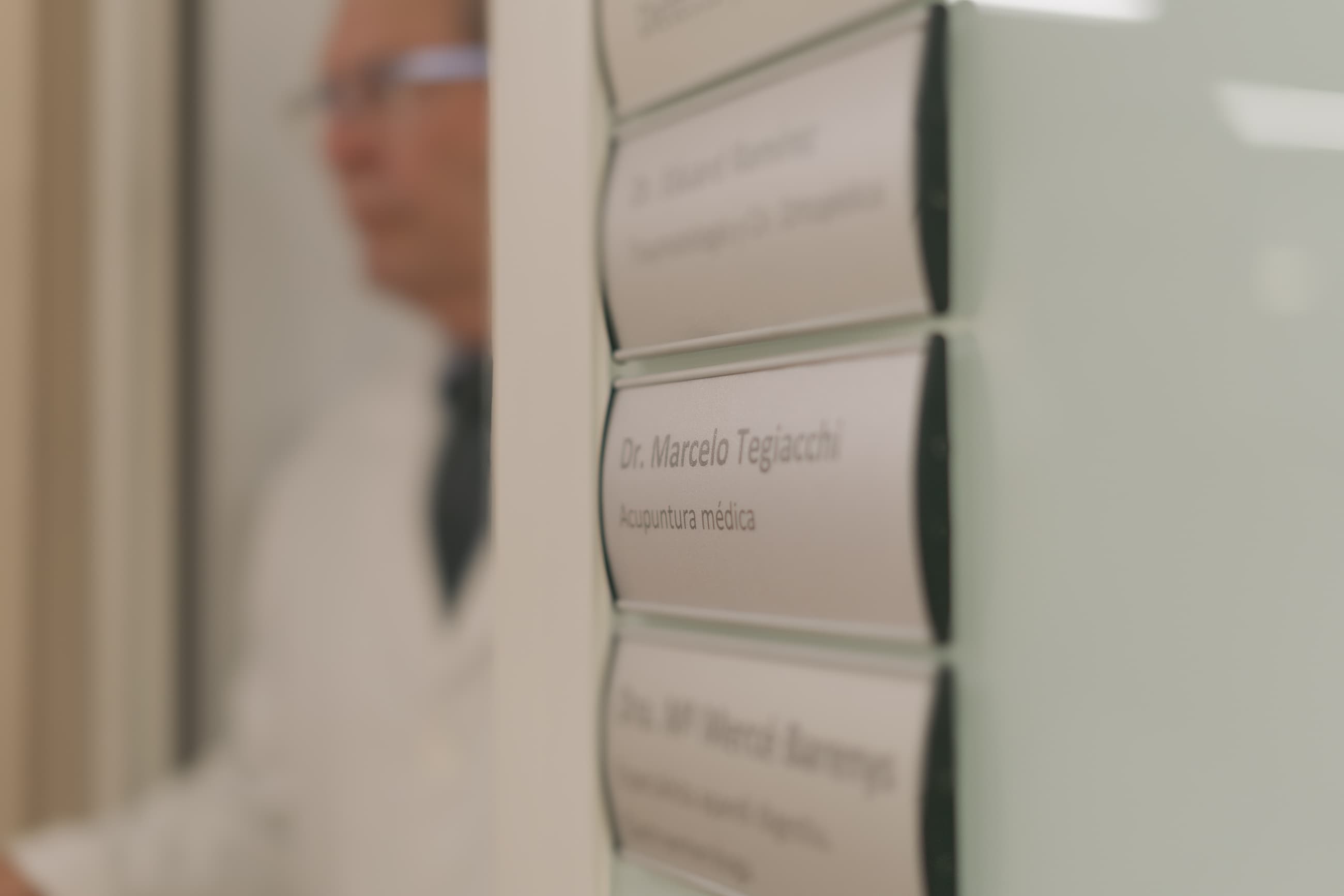 Placa del Dr. Marcelo Tegiacchi, acupuntura médica, con un médico desenfocado en el fondo.