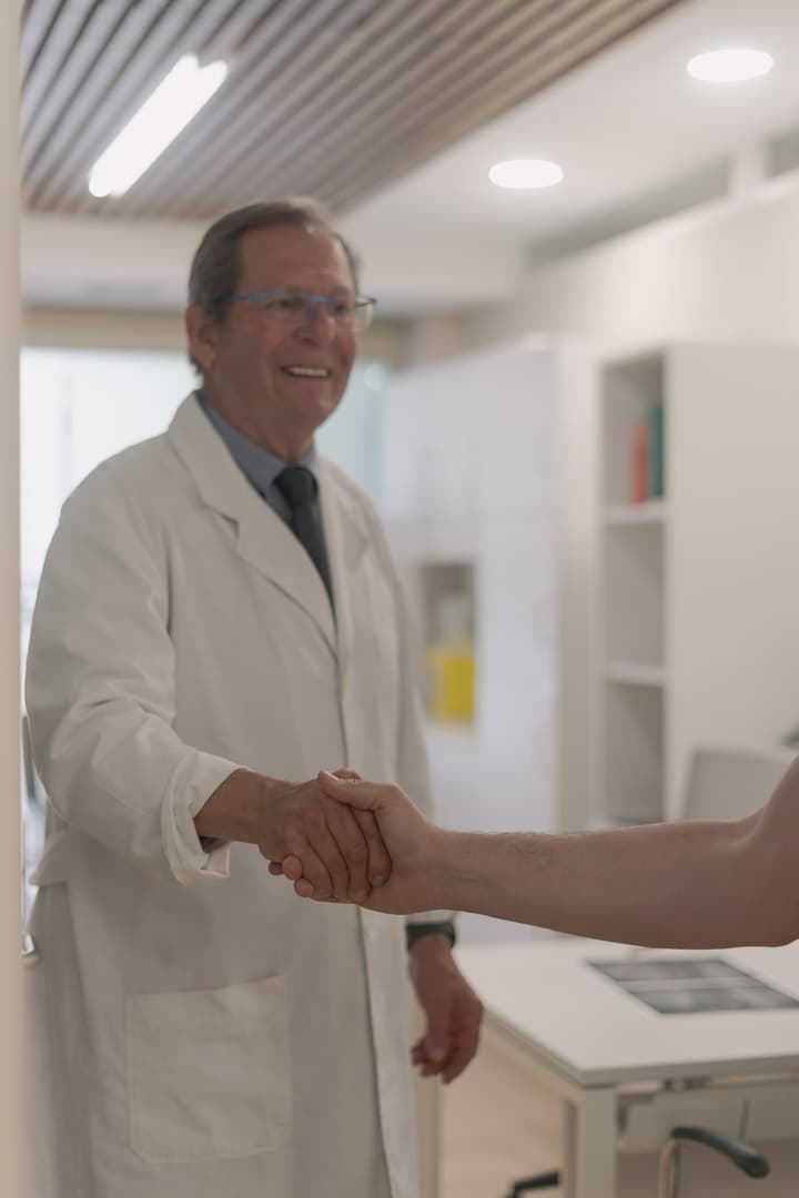 Médico sonriente con bata blanca estrechando la mano de un paciente en un consultorio médico.
