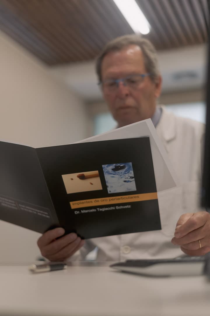 Médico con bata blanca revisando un folleto informativo sobre implantes de oro periarticulares.