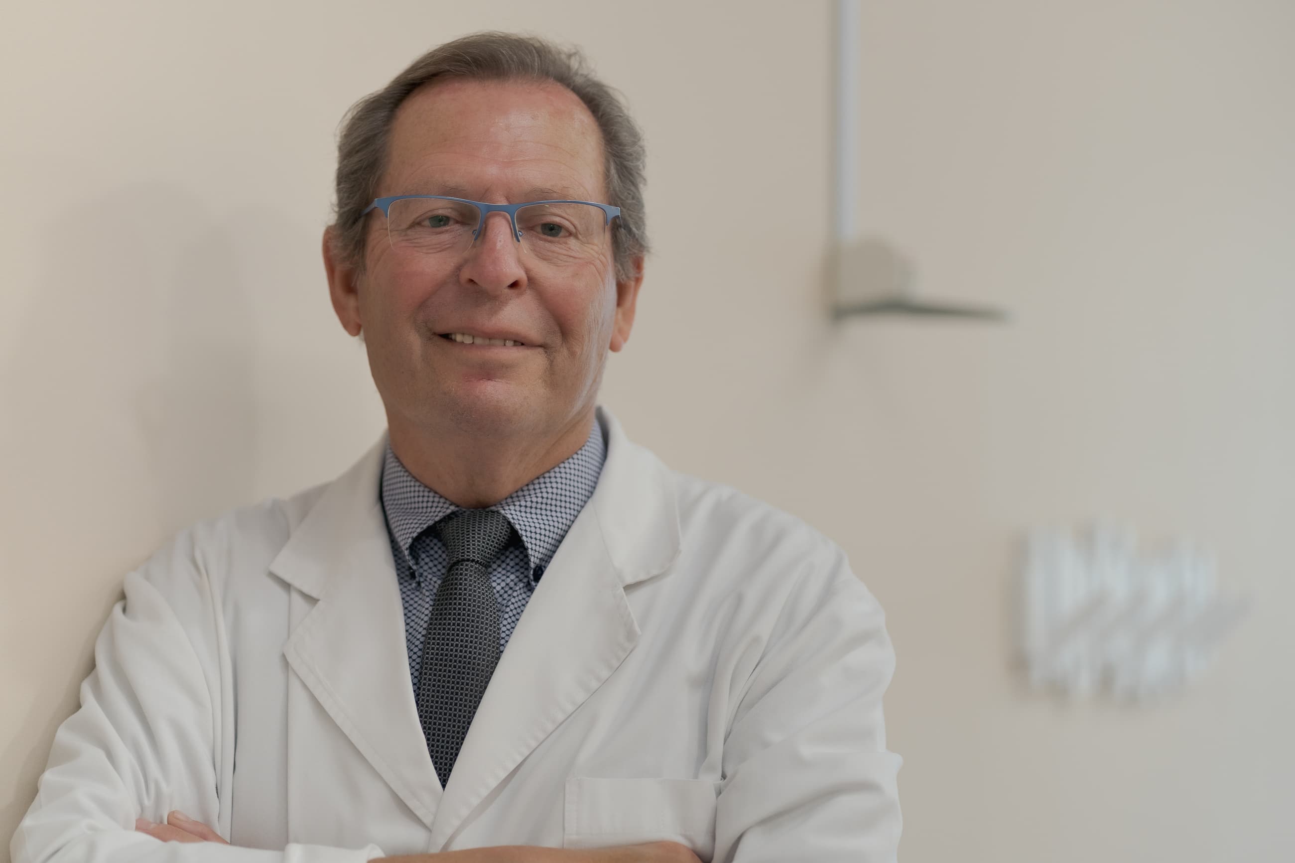 Médico sonriente con bata blanca, gafas azules y corbata gris mirando a la cámara.